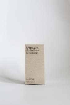 A minimalist-style carton box with typography displaying the words N&eacute;cessaire The Deodorant Le D&eacute;odorant. The text is printed in black against a light beige background. It indicates the eucalyptus scent and a size of 50 ml / 1.7 fl oz. The box stands upright on a flat, light surface with a soft shadow cast beside it.
