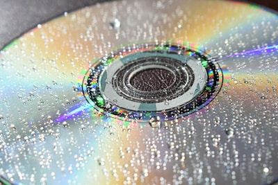 A close-up of a compact disc with numerous water droplets scattered across its surface. The disc reflects a spectrum of colors, creating a vivid and iridescent effect. The water droplets add texture and enhance the play of light on the shiny surface.