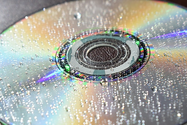 A close-up of a compact disc with numerous water droplets scattered across its surface. The disc reflects a spectrum of colors, creating a vivid and iridescent effect. The water droplets add texture and enhance the play of light on the shiny surface.