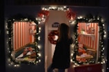 A festive toy shop window decorated with illuminated garlands and red bows. Two display windows reveal toys, including a toy train set, dolls, and miniature vehicles, set against a backdrop of red and white vertical stripes. A person standing outside looks into the windows.