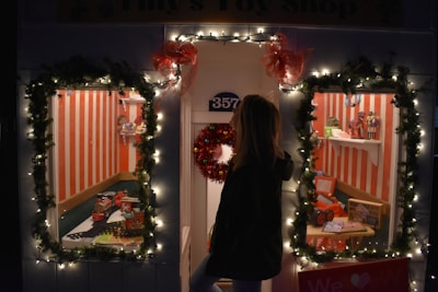 A festive toy shop window decorated with illuminated garlands and red bows. Two display windows reveal toys, including a toy train set, dolls, and miniature vehicles, set against a backdrop of red and white vertical stripes. A person standing outside looks into the windows.