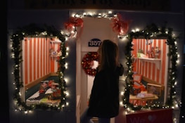 A festive toy shop window decorated with illuminated garlands and red bows. Two display windows reveal toys, including a toy train set, dolls, and miniature vehicles, set against a backdrop of red and white vertical stripes. A person standing outside looks into the windows.