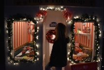 A festive toy shop window decorated with illuminated garlands and red bows. Two display windows reveal toys, including a toy train set, dolls, and miniature vehicles, set against a backdrop of red and white vertical stripes. A person standing outside looks into the windows.