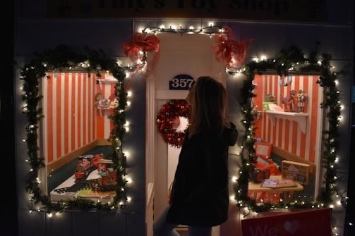 A festive toy shop window decorated with illuminated garlands and red bows. Two display windows reveal toys, including a toy train set, dolls, and miniature vehicles, set against a backdrop of red and white vertical stripes. A person standing outside looks into the windows.