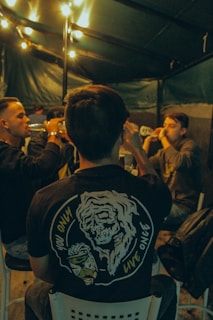 A group of people sitting together, possibly engaged in a casual social gathering. One person has a visible shirt with a skeleton design and the phrase 'You Only Live Once.' The setting appears to be under a canopy with string lights providing warm lighting.