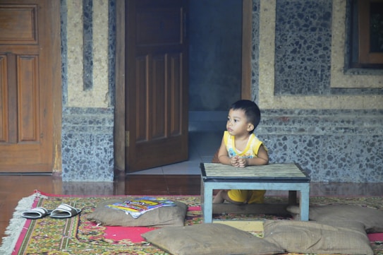A young child wearing a yellow outfit sits on a patterned rug, resting their arms on a small wooden table. Nearby are a pair of sandals and some colorful magazines resting on a cushion. The setting appears to be inside a room with stone walls and wooden doors, creating a cozy and homely atmosphere.