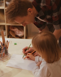 An adult and a child are at a table, engaged in drawing or coloring with pencils. The adult, wearing a plaid shirt, is keenly observing or helping the child, who is focused on the activity. Various colored pencils and other drawing tools are organized in a transparent holder on the table.