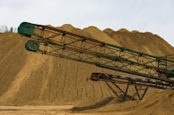 An industrial scene featuring large mounds of sand or gravel with long, rusty green metal conveyors extending across the foreground. The sky is overcast, providing a neutral lighting to the landscape.