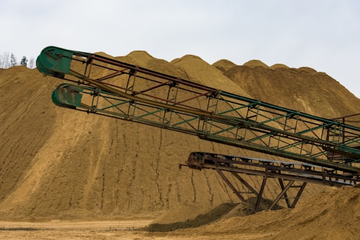 An industrial scene featuring large mounds of sand or gravel with long, rusty green metal conveyors extending across the foreground. The sky is overcast, providing a neutral lighting to the landscape.