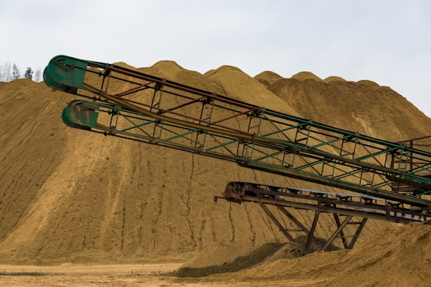 An industrial scene featuring large mounds of sand or gravel with long, rusty green metal conveyors extending across the foreground. The sky is overcast, providing a neutral lighting to the landscape.