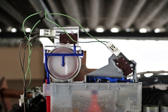 A close-up view of a mechanical or electronic component featuring various parts including wires, connectors, and intricate parts. Several green wires are connected to components on top of a translucent container. The background is blurred, emphasizing the device.
