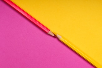 Two colored pencils, one red and one yellow, are aligned on a surface divided diagonally into two contrasting backgrounds, one yellow and one pink. The pencils are positioned point to point, creating a symmetrical and dynamic visual effect.