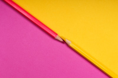 Two colored pencils, one red and one yellow, are aligned on a surface divided diagonally into two contrasting backgrounds, one yellow and one pink. The pencils are positioned point to point, creating a symmetrical and dynamic visual effect.