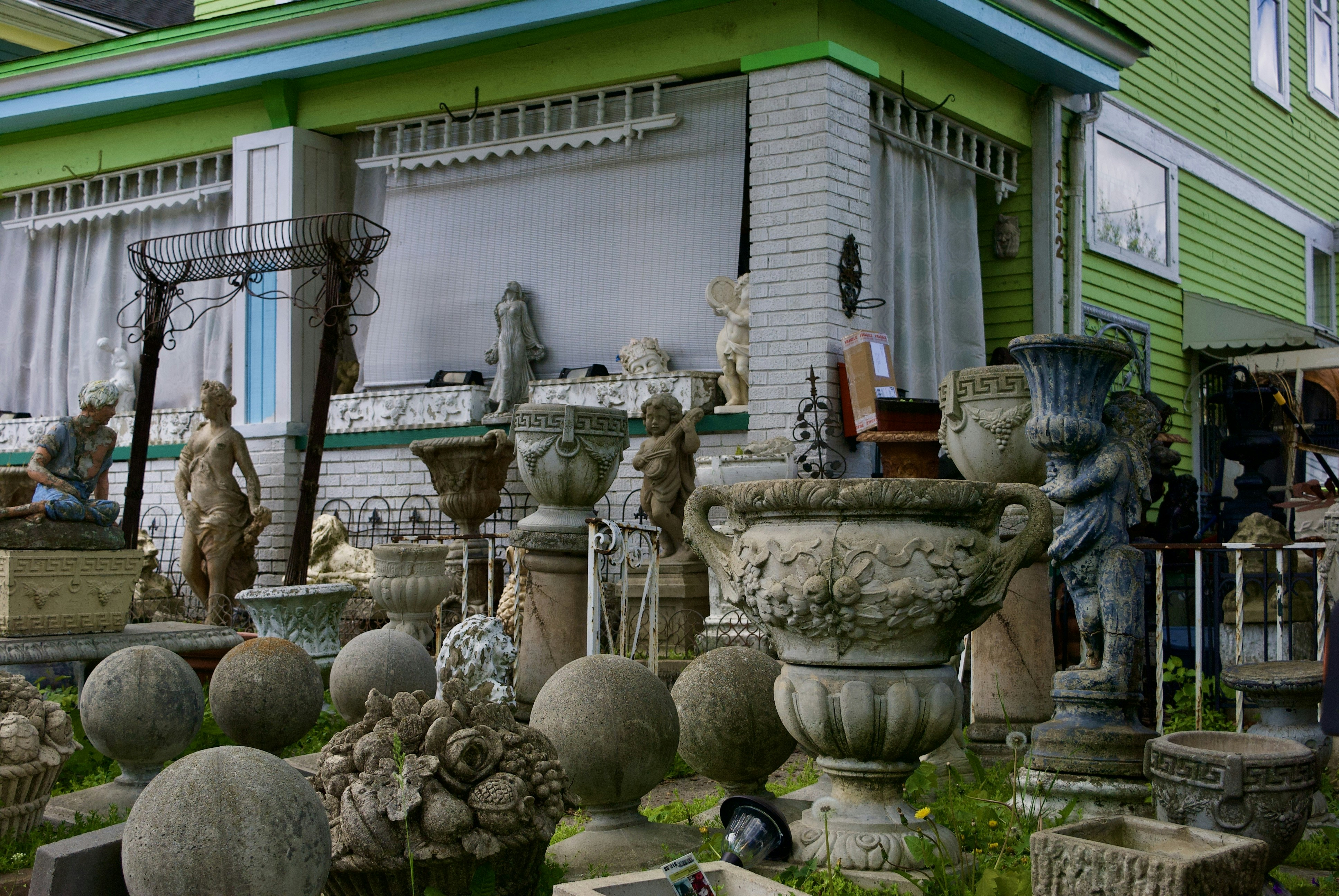 A collection of decorative stone and concrete garden ornaments, including statues, urns, vases, and spherical sculptures, is arranged outside a building with green siding and large windows. The arrangement includes classical and ornate designs, with several human figures and decorative motifs.