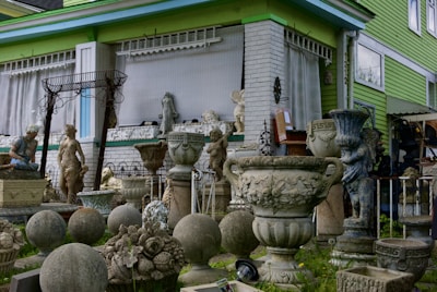 A collection of decorative stone and concrete garden ornaments, including statues, urns, vases, and spherical sculptures, is arranged outside a building with green siding and large windows. The arrangement includes classical and ornate designs, with several human figures and decorative motifs.