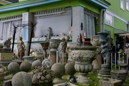 A collection of decorative stone and concrete garden ornaments, including statues, urns, vases, and spherical sculptures, is arranged outside a building with green siding and large windows. The arrangement includes classical and ornate designs, with several human figures and decorative motifs.