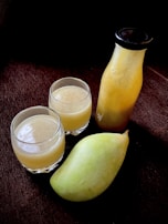 A mango fruit, a bottle filled with mango juice, and two clear glasses containing juice, all placed against a dark background. The juice has a light yellow hue, resembling freshly squeezed mango pulp.