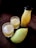 A mango fruit, a bottle filled with mango juice, and two clear glasses containing juice, all placed against a dark background. The juice has a light yellow hue, resembling freshly squeezed mango pulp.