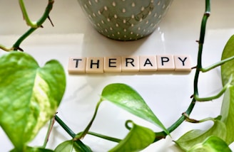 Arranged Scrabble tiles spell out the word 'THERAPY' on a white surface. Bright green leaves frame the image, with a plant pot in the background contributing an organic and serene aesthetic.