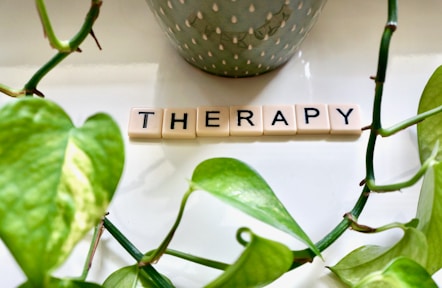 Arranged Scrabble tiles spell out the word 'THERAPY' on a white surface. Bright green leaves frame the image, with a plant pot in the background contributing an organic and serene aesthetic.