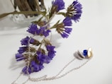 A delicate arrangement featuring dried purple flowers accompanied by a silver chain necklace. The flowers are slightly tilted with a soft-focused background, enhancing their vibrant hue. The necklace, partly coiled, showcases a small gold disc and a blue bead, adding a touch of elegance.