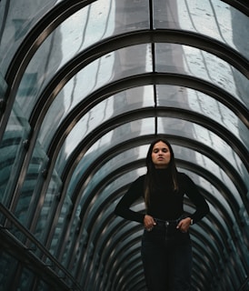 A person stands confidently within a futuristic, arched glass tunnel. The tunnel's reflective surface and metal framework create an intriguing geometric pattern that draws the eye toward the figure at the center.