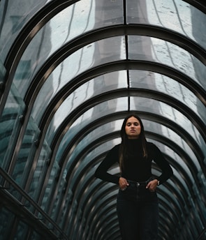 A person stands confidently within a futuristic, arched glass tunnel. The tunnel's reflective surface and metal framework create an intriguing geometric pattern that draws the eye toward the figure at the center.