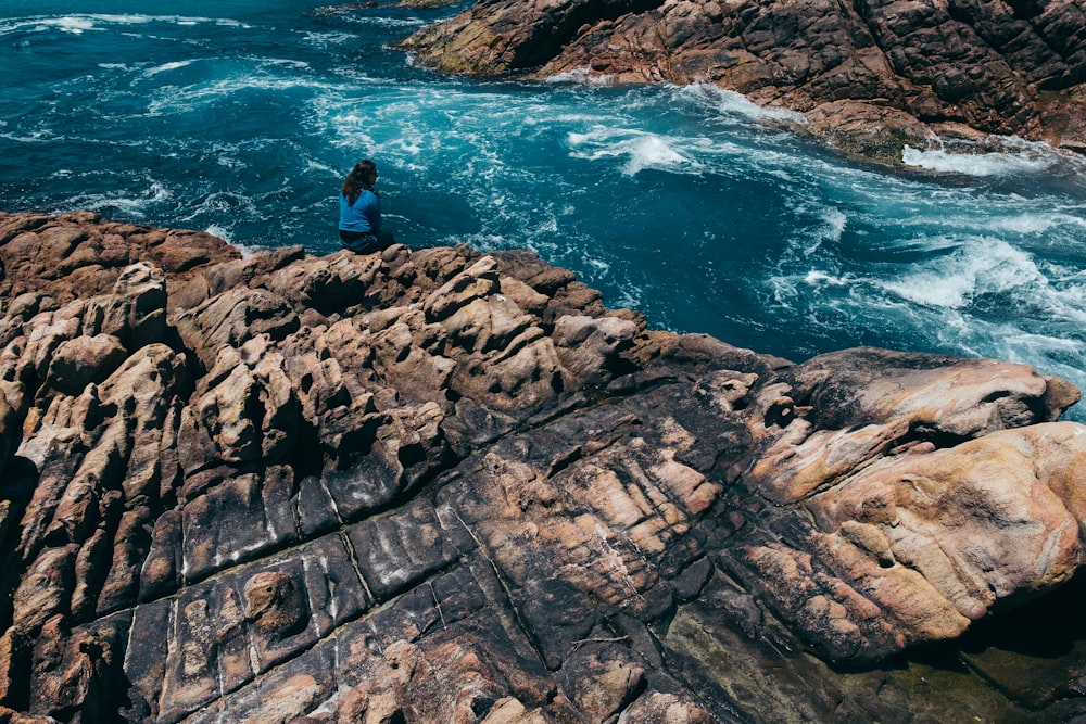 Mann im schwarzen Hemd steht tagsüber auf einer braunen Felsformation in der Nähe eines Gewässers