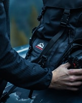 A person is handling a black backpack with a mountain logo and the word 'pursuitretro' on it. The backpack is sitting on a dark surface, possibly a car hood. The person is holding a camera, also visible in the lower right corner. The background is blurred, suggesting an outdoor setting with a subdued, possibly forest environment.