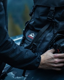 A person is handling a black backpack with a mountain logo and the word 'pursuitretro' on it. The backpack is sitting on a dark surface, possibly a car hood. The person is holding a camera, also visible in the lower right corner. The background is blurred, suggesting an outdoor setting with a subdued, possibly forest environment.