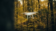 A white quadcopter drone hovers in a forest setting with a blurred background of green and brown trees. The drone's camera is visible, and its propellers are in motion.