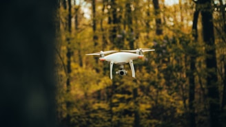 A white quadcopter drone hovers in a forest setting with a blurred background of green and brown trees. The drone's camera is visible, and its propellers are in motion.