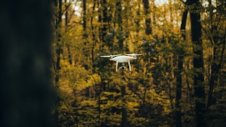 A white drone hovers mid-air in a forest with dense trees and foliage surrounding it. The background is filled with shades of green and brown, giving an autumnal feel.