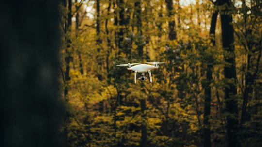 A white drone hovers mid-air in a forest with dense trees and foliage surrounding it. The background is filled with shades of green and brown, giving an autumnal feel.