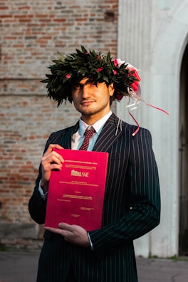 A person in a suit and tie holds a red academic diploma or certificate. They are wearing a laurel wreath decorated with red and white ribbons. The background features a textured brick wall.