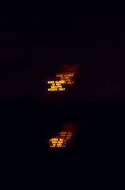 A dimly lit scene with an illuminated road sign displaying a closure notice in bright orange text. The sign is partially blurred, likely due to motion, and its reflection can be seen below in a darker area.