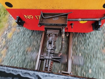 The image shows a close-up view of a train coupling mechanism, featuring metallic components connected to railway tracks. The background consists of blurred gravel and tracks, indicating movement.
