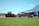 A large red agricultural tractor with tracks, connected to an equally large planting machine, is positioned near a barn against a backdrop of open fields and a clear blue sky. The barn appears white and the grass is neatly manicured.