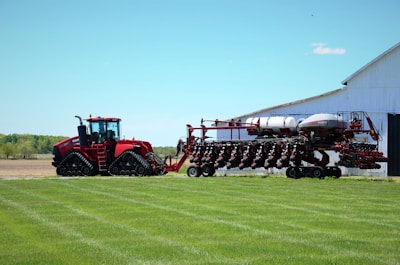 A large red agricultural tractor with tracks, connected to an equally large planting machine, is positioned near a barn against a backdrop of open fields and a clear blue sky. The barn appears white and the grass is neatly manicured.