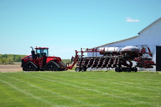 A large red agricultural tractor with tracks, connected to an equally large planting machine, is positioned near a barn against a backdrop of open fields and a clear blue sky. The barn appears white and the grass is neatly manicured.