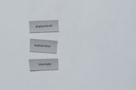 Three pieces of paper arranged vertically feature the words 'dialectical', 'behaviour', and 'therapy'. The background is a plain light gray.