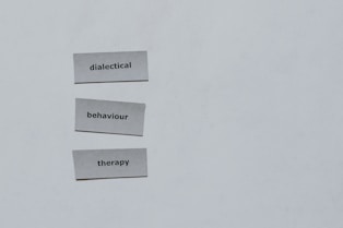 Three pieces of paper arranged vertically feature the words 'dialectical', 'behaviour', and 'therapy'. The background is a plain light gray.