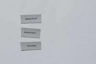 Three pieces of paper arranged vertically feature the words 'dialectical', 'behaviour', and 'therapy'. The background is a plain light gray.