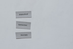 Three pieces of paper arranged vertically feature the words 'dialectical', 'behaviour', and 'therapy'. The background is a plain light gray.