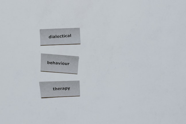 Three pieces of paper arranged vertically feature the words 'dialectical', 'behaviour', and 'therapy'. The background is a plain light gray.