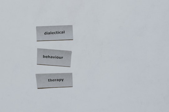Three pieces of paper arranged vertically feature the words 'dialectical', 'behaviour', and 'therapy'. The background is a plain light gray.
