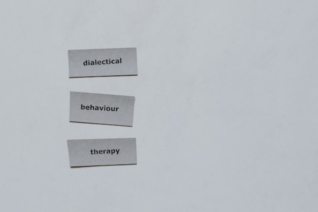Three pieces of paper arranged vertically feature the words 'dialectical', 'behaviour', and 'therapy'. The background is a plain light gray.