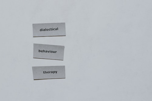 Three pieces of paper arranged vertically feature the words 'dialectical', 'behaviour', and 'therapy'. The background is a plain light gray.