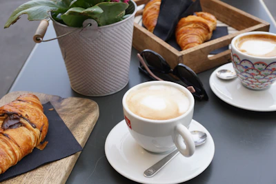 An outdoor table setting with a copy of one of the books beside a cup of espresso and a croissant.