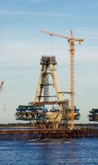 A partially constructed bridge visible over a body of water, featuring a prominent A-shaped structure being supported by cranes. The construction is ongoing with scaffolding and machinery, surrounded by a clear sky.